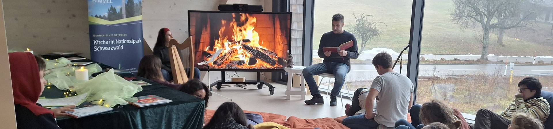 Mann liest einer Gruppe vor, im TV ist ein Lagerfeuer zu sehen